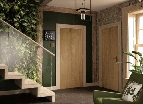 A wooden staircase ascends beside a lush vertical garden wall, leading to a hallway with two wooden doors. A green armchair and a stone wall are illuminated by natural light.